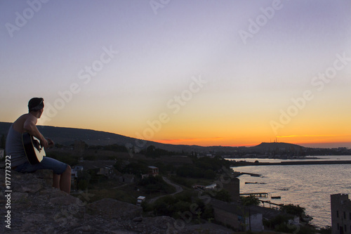 man with guitar sits on a hill in the background of sunset. see the sea