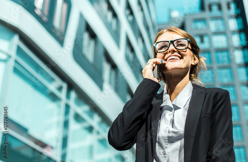 Young blonde caucasian businesswoman in a white blouse and a black jacket is talking phone before the modern corporation building in the background