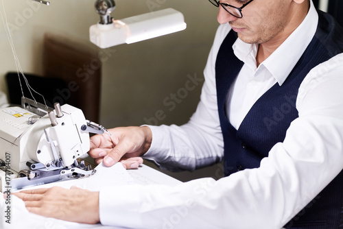 Portrait of handsome tailor sewing clothes at table in atelier working with  serger machine in dim light photo