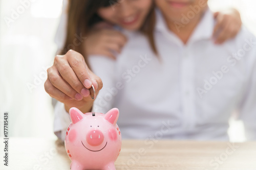Smile couple putting a coin into a pink piggy bank on wooden desk - save money for the future.