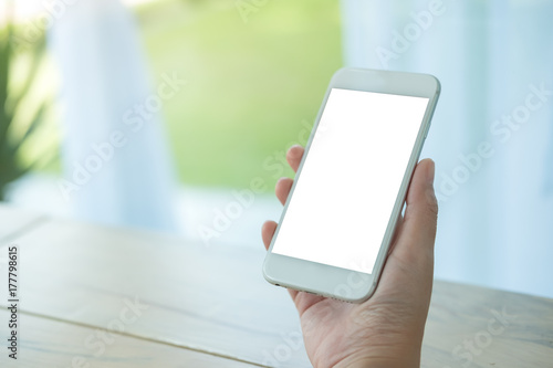 Mockup image of hand holding white mobile phone with blank screen on table in cafe