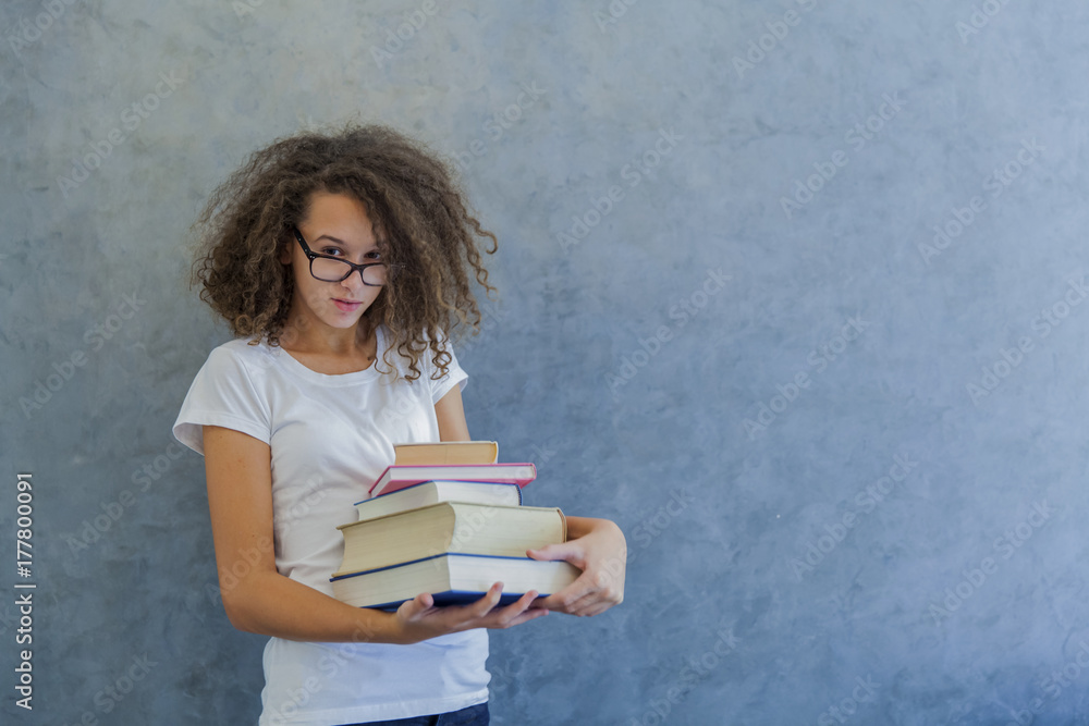 Curly hair teen girl with glasses stands next to the wall and holds several books Curly hair teen girl with glasses stands next to the wall and holds several books