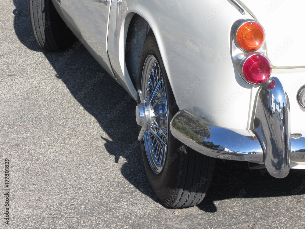 Rear of an old british classic car . Particular view of left tail light, shiny chrome bumper and left rear tire . The car is a Triumph TR3 model produced between 1955 and 1962