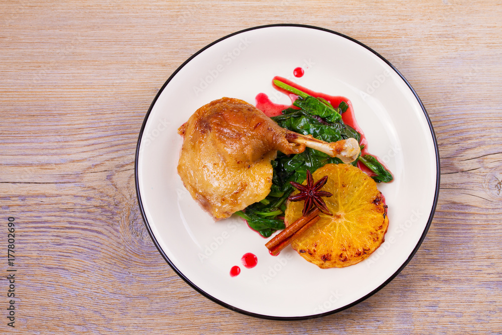 Duck leg with spinach and orange in sweet and sour berry sauce, white plate on rustic wooden table Duck leg with spinach and orange in sweet and sour berry sauce, white plate on rustic wooden table