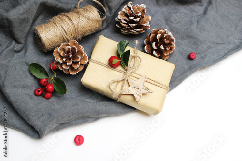 Christmas festive styled stock image composition. Handmade Christmas gift box, red berries, pine cones isolated on grey linen blanket. Flat lay, top view. Winter holiday background. Winter concept.