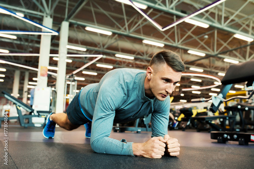 Portrait of fit young man doing plank excursive on floor in modern gym, looking away with determination