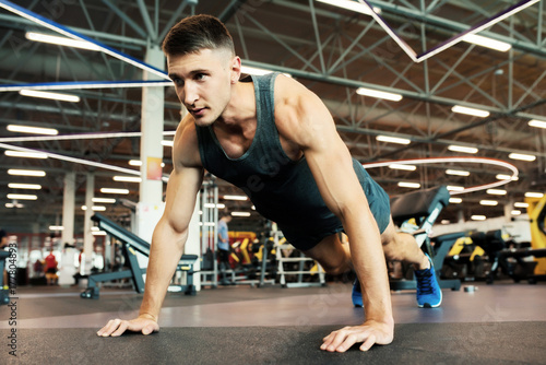 Portrait of handsome young man with muscular arms doing push ups on floor in modern gym