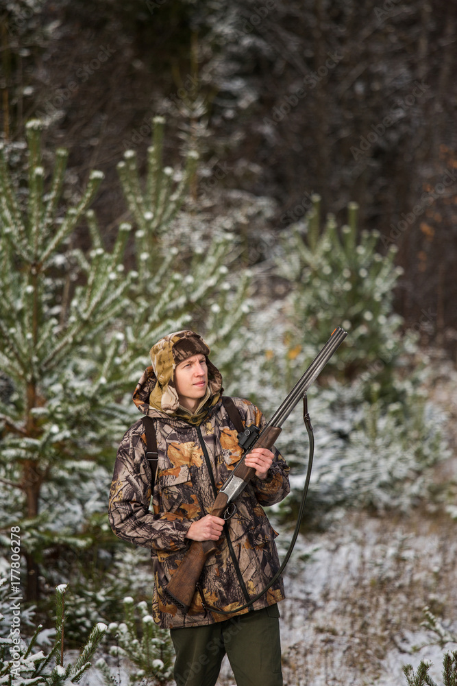 Male hunter with backpack, armed with a rifle, standing in a snowy winter forest. Man looks away. Snowfall in the forest. Male hunter with backpack, armed with a rifle, standing in a snowy winter forest. Man looks away. Snowfall in the forest.