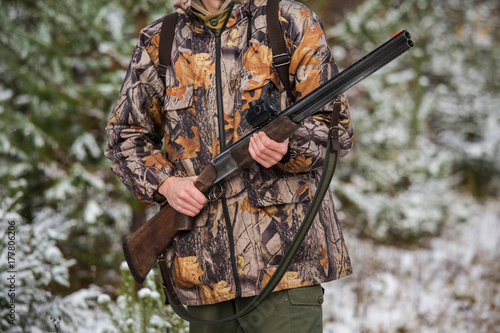 Male hunter in camouflage, armed with a rifle, standing in a snowy winter forest.