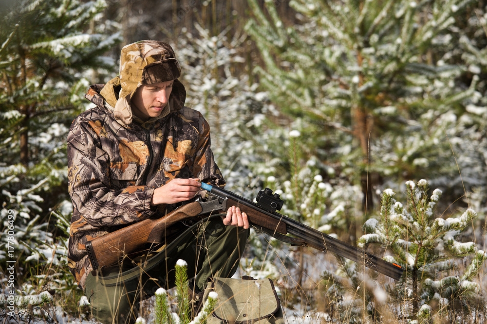 Hunter with a backpack and a hunting gun in the winter forest. Man is charging a hunting rifle. Winter snow-covered forest.