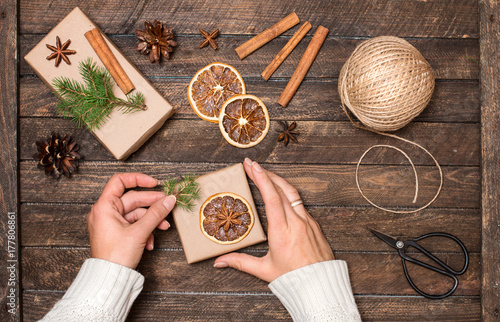 Woman decorating Christmas gifts. Presents wrapping inspirations. Hands, gift boxes, ball of jute, cinnamon sticks, anise, orange slices, fer tree branches and retro scissors.