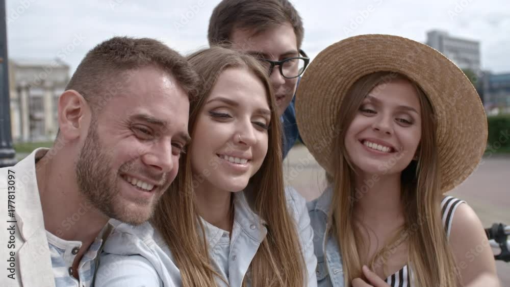 Closeup of young happy women and men sitting together outdoors, smiling and posing at camera while taking a selfie with smartphone Closeup of young happy women and men sitting together outdoors, smiling and posing at camera while taking a selfie with smartphone