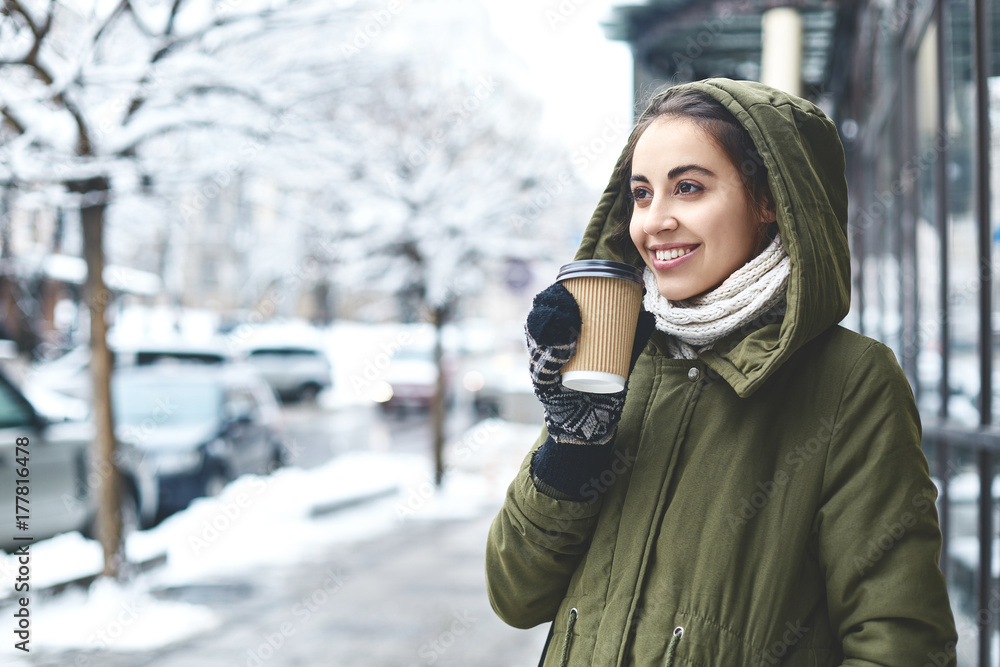 young beautiful brunette woman in a warm jacket with hoodie and warm mittens is walking outdoors in winter. a woman holding a paper glass with hot tea or coffee. Enjoys a walk on a frosty day young beautiful brunette woman in a warm jacket with hoodie and warm mittens is walking outdoors in winter. a woman holding a paper glass with hot tea or coffee. Enjoys a walk on a frosty day