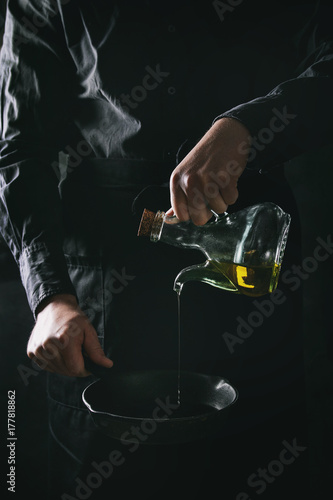 Man chef in black apron pouring olive oil from bottle for cooking pancakes in cast-iron pan. Dark rustic style. Toned image