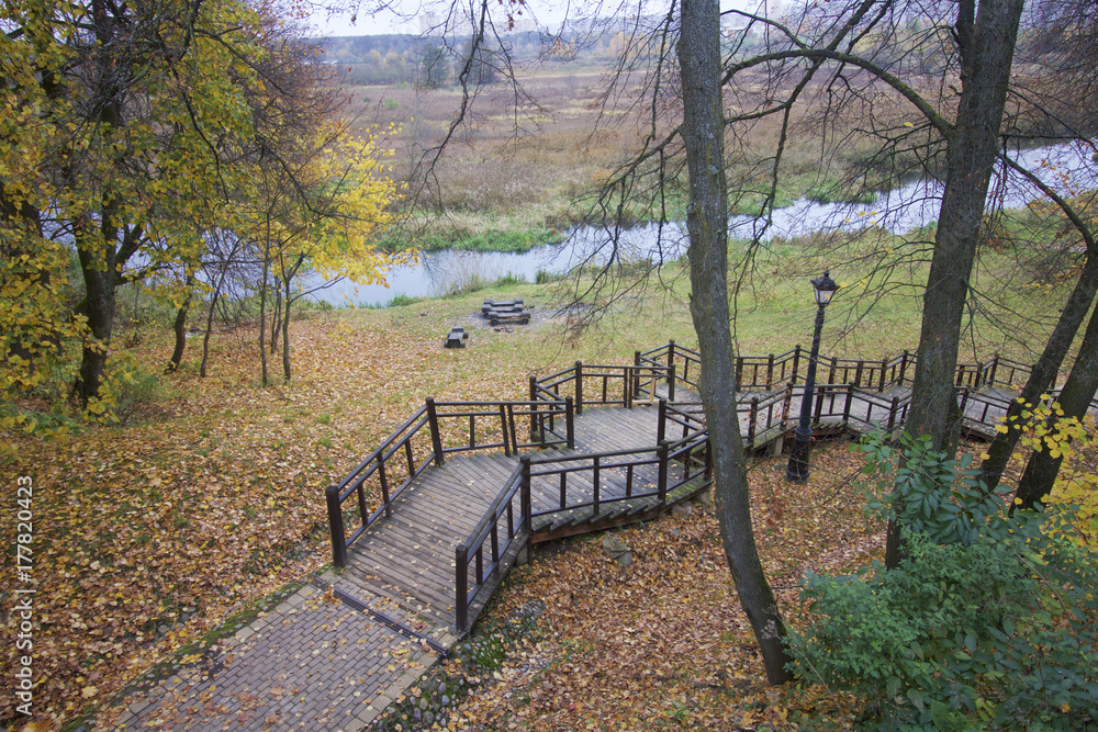 Autumn landscape in the city park. On the ground a carpet of fallen yellow leaves of different shades. From the pedestrian bridge you can see wooden steps coming down the slope towards the river.