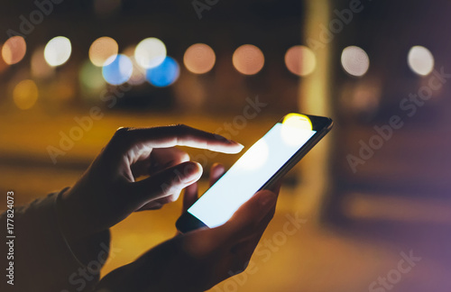 Woman pointing finger on screen smartphone on background bokeh light in night atmospheric city, hipster using in hands mobile phone closeup, mockup glitter street, online wifi internet concept