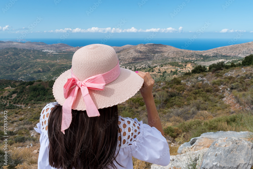 Beautiful girl with a wide hat, standing with her back on a background of green mountains and the sea