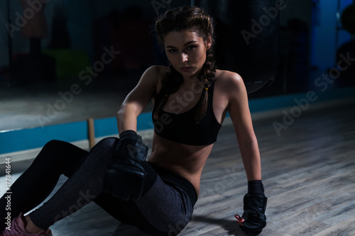 Young fighter boxer girl wearing boxing gloves sitting on the floor of dark gym before  training.