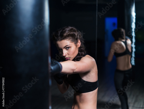 Cute fighter boxer fit girl wearing boxing gloves in training with heavy punching bag in gym. She is in good shape. Model looking at camera