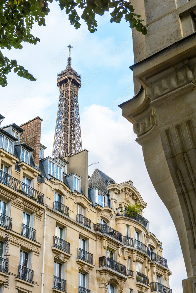 The top of the Eiffel Tower seen from down the street with foliage and typical residential buildings in the foreground.