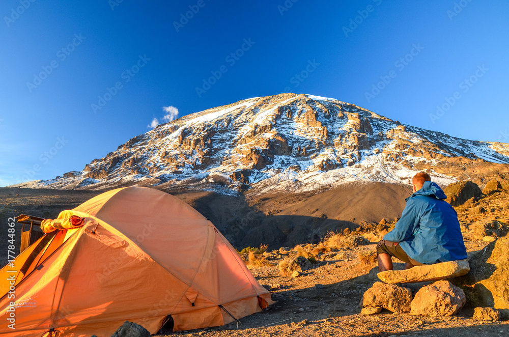 Fototapeta premium Wieczorny widok na Kibo ze szczytem Uhuru (5895 m npm, najwyższa góra w Afryce) na Kilimandżaro w Parku Narodowym Kilimandżaro, widziany z obozu Karanga na 3995 m npm. Namiot i młody turysta na pierwszym planie.