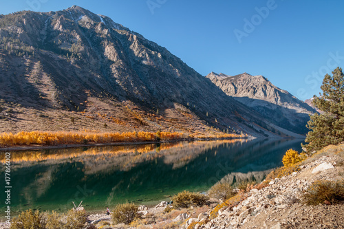 Lake in the Eastern Sierra Nevada Mountains in the fall color. Aspen line the lake with high cliffs and blue sky in the background. There are people fishing around the lake.