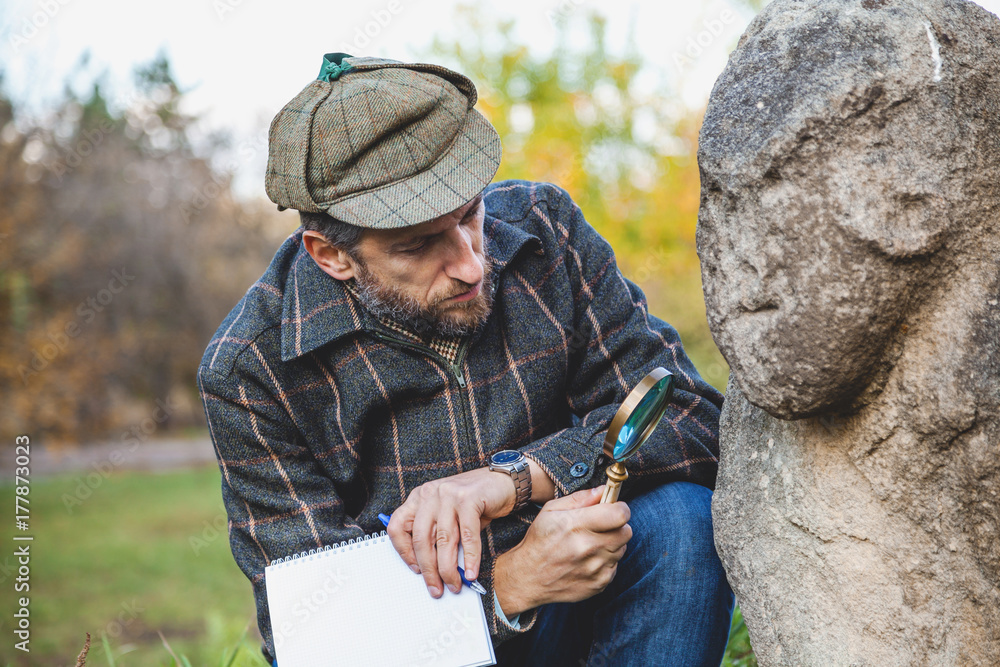 Scientific historian examines through magnifying glass stone sculpture on mound