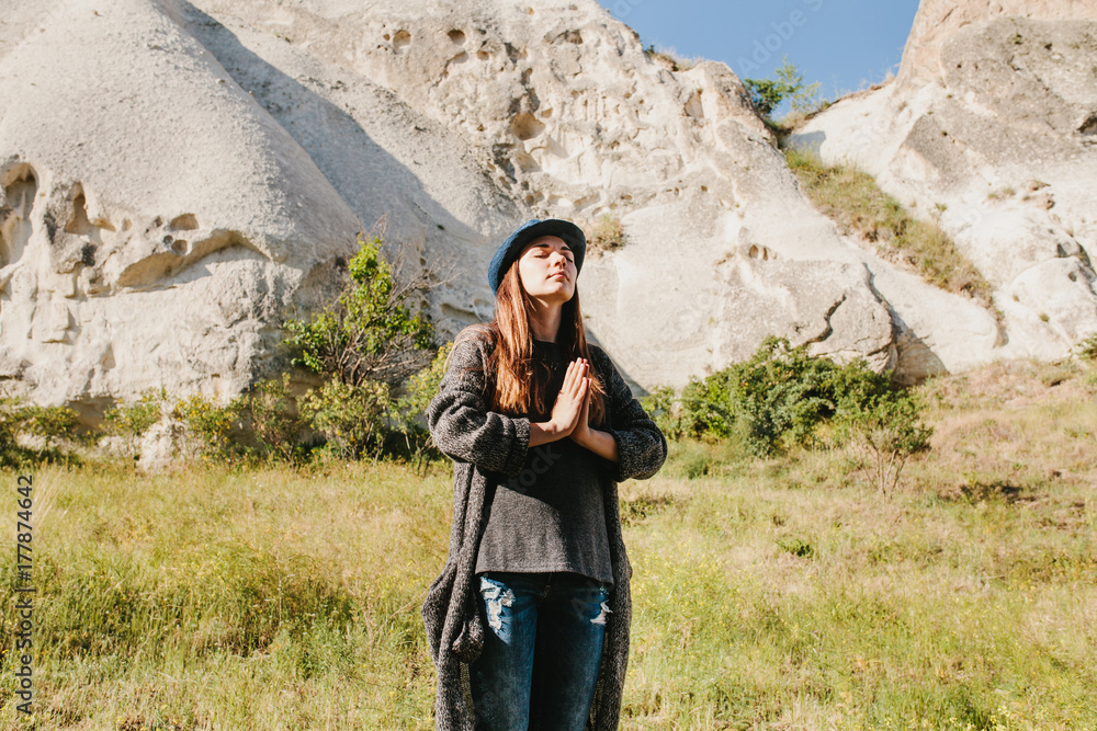 Naklejka premium Beautiful young woman in hat and gray sweater meditates with folded hands and closing eyes against backdrop of beautiful mountains and hills on sunny day