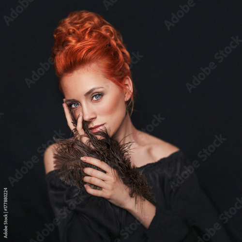 Portrait of redhead woman with blue eyes wearing black dress on dark background. Girl looks directly into the camera.