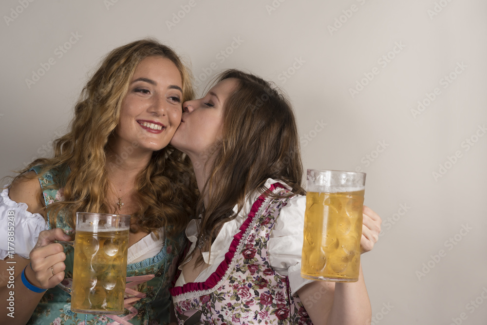 Girl in Dirndl Kisses Another on Cheek Holding Mugs of Beer Girl in Dirndl Kisses Another on Cheek Holding Mugs of Beer