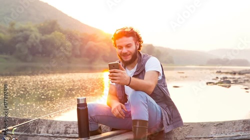 Stylish beautiful fishman talking on a smartphone on a wooden boat. The man speaks on Skype at sunset.