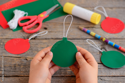 Child made red and green Christmas balls from cardboard paper. Child holds Christmas ball in his hands. Step. Stationery on the wooden table. Easy Christmas crafts for kids to make 