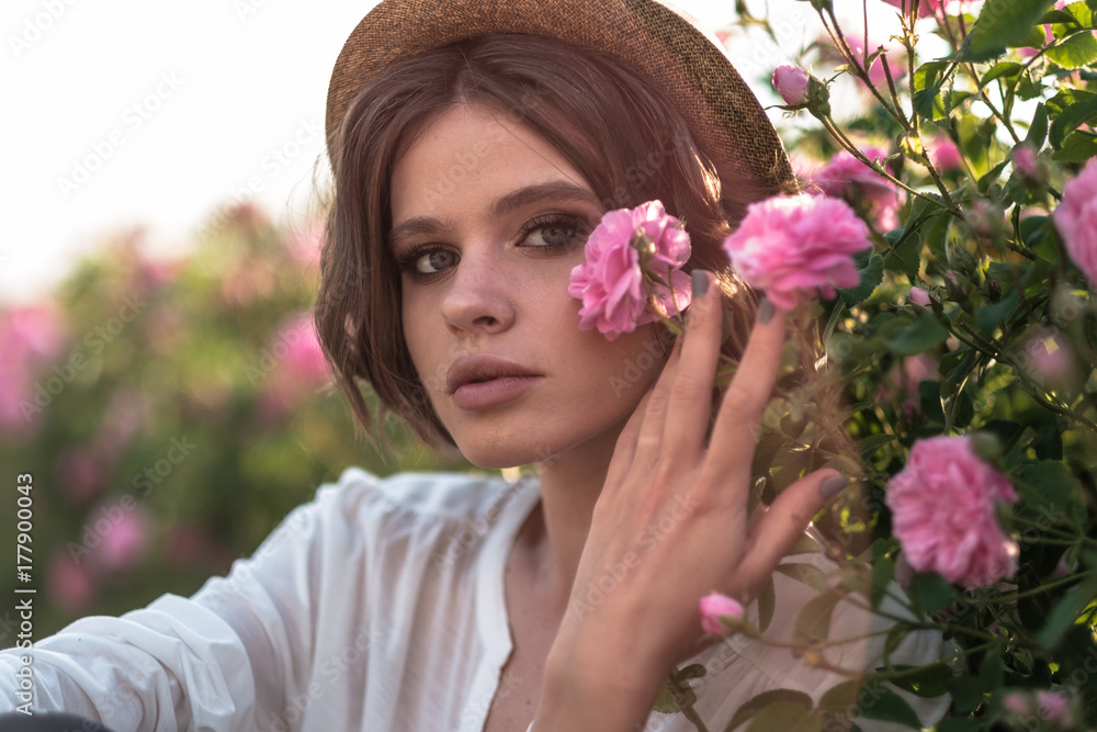 Fototapeta premium Beautiful young woman with curly hair posing near roses in a garden. The concept of perfume advertising.