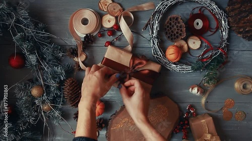 A top-down plan. Fully visible the table with the decorations. Female hands put and finalize Christmas gift wrapped in craftool paper on a wooden table. Bandaging tape and tied in a bow. photo