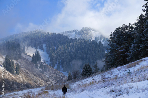 Bild auf Leinwand Man stands affront massive mountains in snow and forest under sky with low white clouds