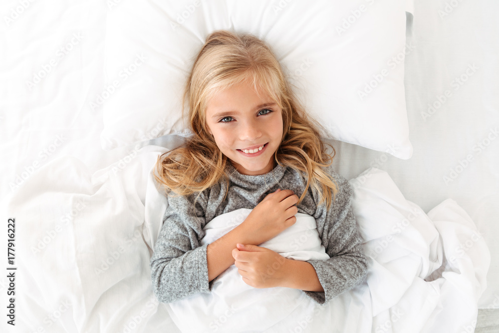 Top view of charming blonde girl smiling and looking at camera while lying in bed Top view of charming blonde girl smiling and looking at camera while lying in bed