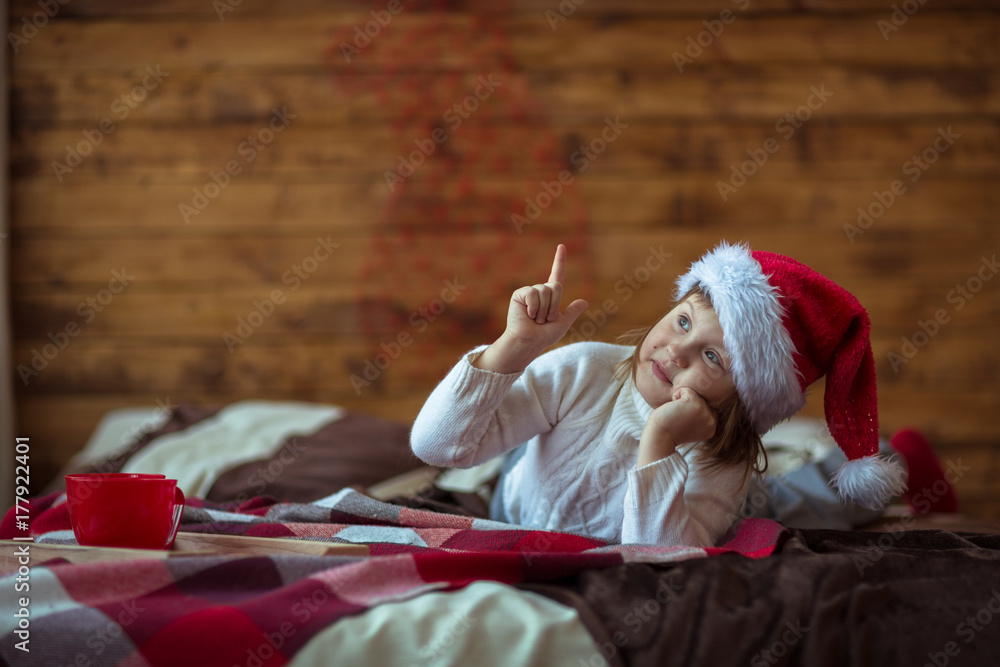 Obraz premium Girl child in jersey in santa hat on the bed, lifestyle, real interior, rustic style, red and brown wooden background