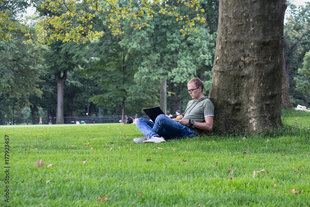 Young man in glasses work on laptop sitting on the grass near the tree in park. Maybe student or hacker businessman Young man in glasses work on laptop sitting on the grass near the tree in park. Maybe student or hacker businessman