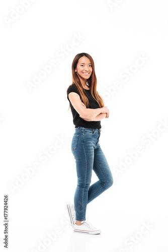Side view of young pretty chinese woman in black tshirt and jeans standing with crossed hands, looking at camera