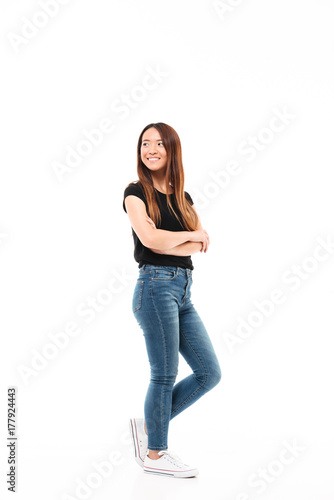 Full length photo of young pretty chinese woman in black tshirt and jeans standing with crossed hands, looking aside