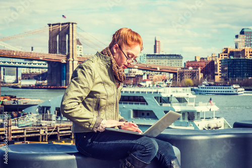 American business woman traveling, working in New York, wearing green leather jacket, jeans, scarf, glasses, sitting by East River, working on laptop computer. Brooklyn bridge, boat on background.. © Alexander Image