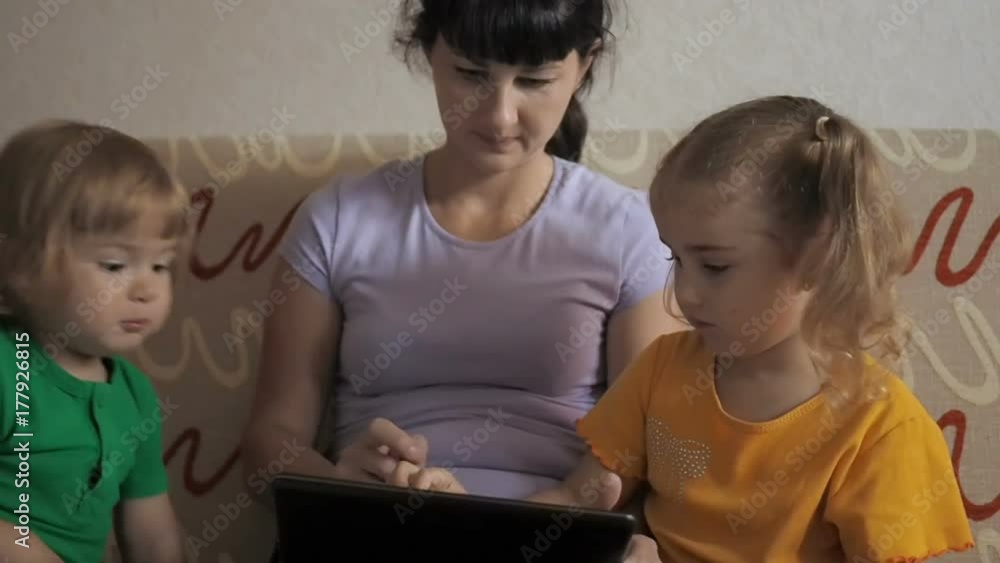 Pretty little girl, boy and mother sitting on sofa using laptop. An adorable family sit together and play with a laptop and digital tablet. Pretty little girl, boy and mother sitting on sofa using laptop. An adorable family sit together and play with a laptop and digital tablet.