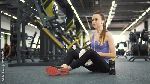 Smiling caucasian girl, in pink vest and white headphones, having water during rest at the gym photo