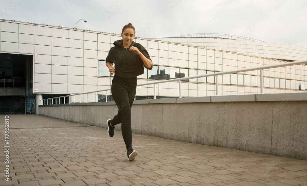 Young attractive lady jogging in the city near buildings. Beautiful happy girl running in the morning outside. Woman trains during the sunrise in the city center before breakfast.	