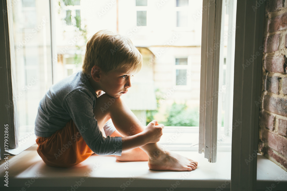 Sad preschooler boy sitting on windowshill. A child looks out the window. Vintage color. Childhood, relocation loneliness concept. Sad preschooler boy sitting on windowshill. A child looks out the window. Vintage color. Childhood, relocation loneliness concept.