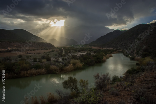 Shafts of sunlight shine through the clouds at sunset over a river in dry countryside