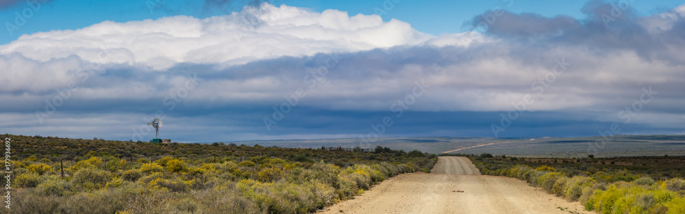 Panoramic Karoo scene in South Africa, a dirt road stretches to the horizon with a windmill pump in the background