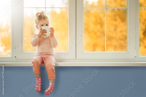 Little cute Kid Girl sitting by window holding cup of hot drink cocoa enjoying autumn forest background. Season Beauty fashion Children concepts. Fall season