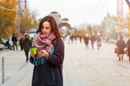 young beautiful brunette girl with  coffee to go  and smartphone on the background of blurred city  street