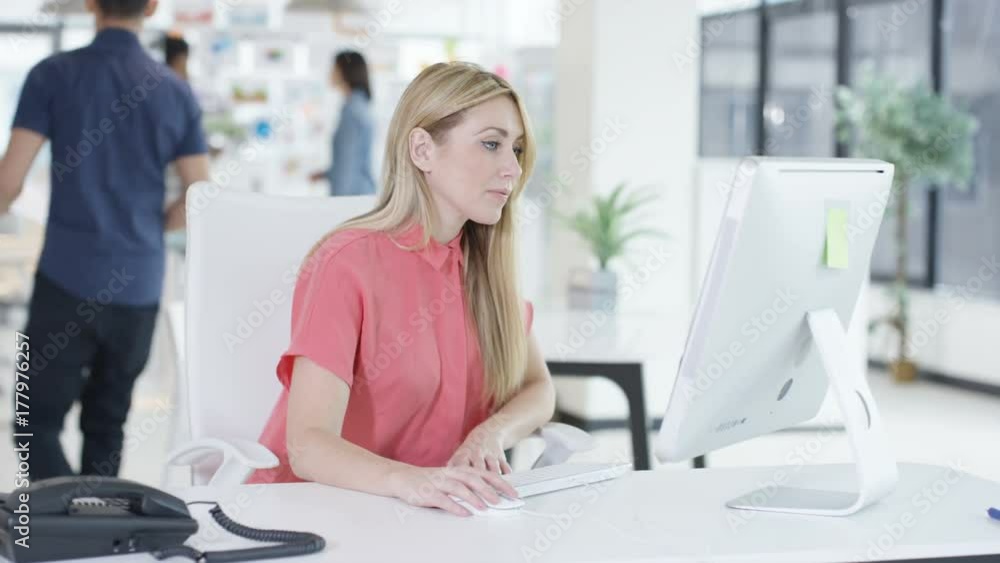 Portrait smiling woman working on computer in creative office with colleagues talking in background. Shot on RED Epic. Portrait smiling woman working on computer in creative office with colleagues talking in background. Shot on RED Epic.